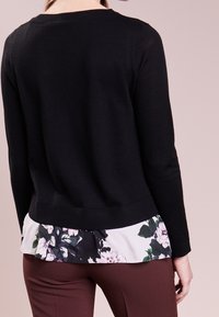Černý svetr s žebrovaným lemem, vrstvený přes blůzu s květinovým vzorem v tónech černé, zelené a fialové. Jednoduchý pohled zezadu.