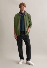 Zielona dzianinowa kurtka zapinana na zamek, na granatowej koszuli, w połączeniu z czarnymi dżinsami i białymi trampkami. Szalik w wzory wokół szyi.