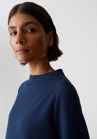 Femme aux cheveux courts et foncés portant un haut texturé bleu marine, regardant légèrement sur le côté devant un fond clair.