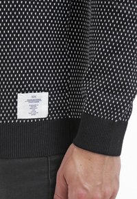 Pull en tricot noir avec un motif texturé de petites formes blanches. Comprend une étiquette en tissu blanc avec la marque et les instructions d'entretien.
