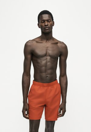 Slank mand med kort krøllet hår iført orange badebukser, stående med lukkede øjne mod en ensartet lys baggrund.