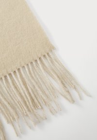 Beige sjaal met een textured breipatroon en franjes aan de uiteinden, gemaakt van een zachte, lichte stof die geschikt is om te lagen.