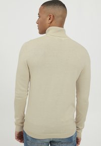 Bež turtleneck pulover z rebrasto teksturo in dolgimi rokavi, dopolnjen s prilegajočo se silhueto. Priložnostno oblačenje brez vidnih vzorcev.