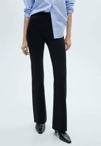 Black flared trousers made of stretchy fabric, featuring a high waistband and a slight split at the hem, paired with a light blue striped shirt.