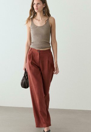 Donna che cammina indossando un top beige senza maniche a coste, pantaloni ampi a vita alta color ruggine, con una piccola borsa marrone scuro.