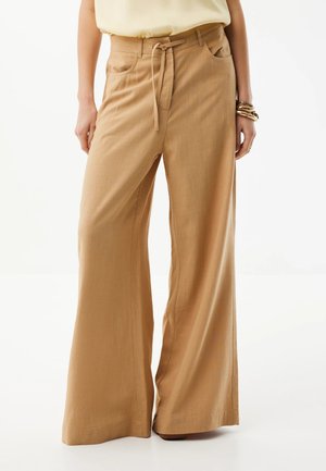 Kobieta ubrana w beżowe, szerokie spodnie z wysokim stanem, z przednimi kieszeniami i paskiem zawiązywanym na supeł, w połączeniu z jasnym topem i bransoletkami.