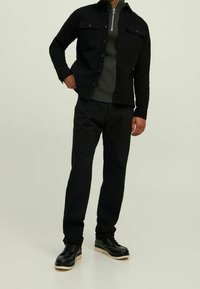 Uomo che indossa una giacca nera con bottoni, un maglione grigio scuro a coste con mezze zip, pantaloni neri e scarpe di pelle nere con suole bianche.