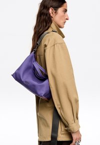 Violetti nailonilaukku, jolla on väljähkö muoto, varustettuna kahdella yläkahvalla ja irrotettavalla mustalla hihnalla, esillä neutraalilla taustalla.