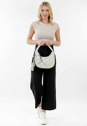Sac à main blanc avec des rayures noires diagonales, surface texturée, anse supérieure arrondie et accent de pompon. Le mannequin porte un haut ajusté et un pantalon large.