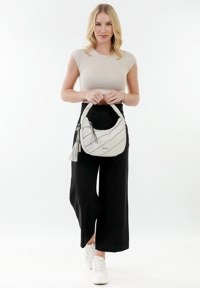 Witte handtas met diagonale zwarte strepen, gestructureerd oppervlakte, afgeronde bovenhandgreep en een franjesaccent. Model draagt een aansluitend topje en wijde pantalon.