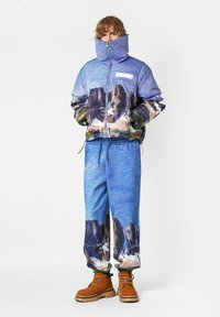 Chaqueta azul con estampado y cuello alto con cremallera, combinada con pantalones deportivos a juego. Botas marrones completan el conjunto. Tela texturizada en toda la prenda.