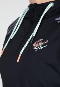 Zwarte pullover met een kwart-rits, voorzien van teal en zwarte getailleerde trekkoordjes, en een koraal logo op de borst. Gladde textuur.