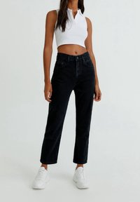 Femme portant un jean noir à taille haute et coupe droite, un haut blanc sans manches court, et des baskets blanches, debout devant un fond clair uni.