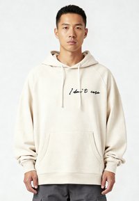 Felpa beige con cappuccio e tasca frontale, con testo scritto a mano nero "Non mi interessa" sul petto. Tessuto morbido, maniche raglan.