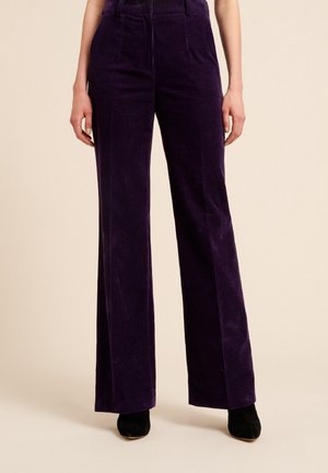 Femme portant un pantalon en velours côtelé large et taille haute de couleur violet foncé avec des chaussures à talons noires, se tenant contre un fond beige uni.
