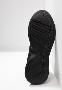 Semelle de chaussure de sport noire en caoutchouc texturé, avec des rainures inclinées et une zone lisse au niveau des orteils. Logo embossé sur le côté.