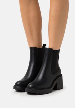 Botines negros con tacón grueso, hechos de cuero suave. Cuentan con paneles laterales elásticos y una suela texturizada con un patrón en zigzag.