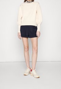 Cremefarbener Pullover mit Puffärmeln, kombiniert mit navyblauen Shorts mit goldenen Knöpfen. Weiße und rosa Sneakers runden das Outfit ab.