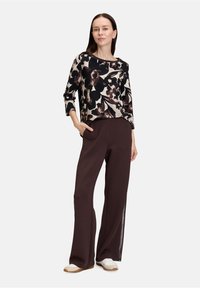 Schwarz-beige Oberteil mit floralem Muster und 3/4-Ärmeln, kombiniert mit dunkelbraunen weiten Hosen. Weiße Sneakers runden das Outfit ab.