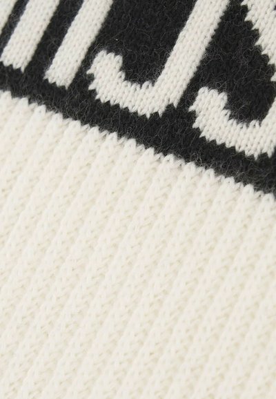 Beanie in maglia di colore crema con testo nero; presenta una texture a coste e un motivo diagonale. Materiale morbido e caldo, adatto per l'uso in inverno.