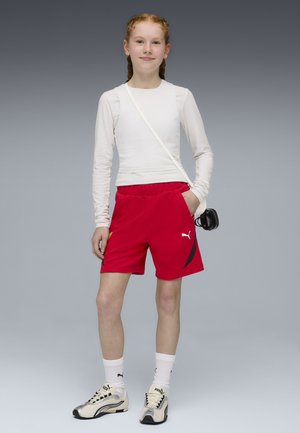 Jeune personne debout, les mains dans les poches, portant une chemise blanche à manches longues, un short rouge Puma, des chaussettes blanches et des baskets beige Puma.