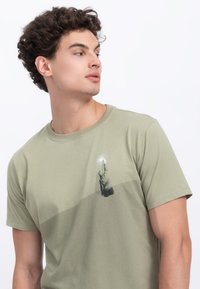 T-shirt in cotone verde oliva con una grafica di una figura che raggiunge verso l'alto, con un effetto sfumato sul tessuto.