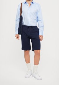 Man som bär en ljusblå skjorta med knappar, marinblå shorts, vita sneakers, vita strumpor och bär en marinblå tygväska med bruna remmar.