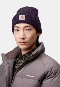 Gorro morado oscuro acanalado con un parche de logo cuadrado. Chaqueta acolchada gris con cuello alto, usada sobre una camisa de franela con patrón a cuadros.