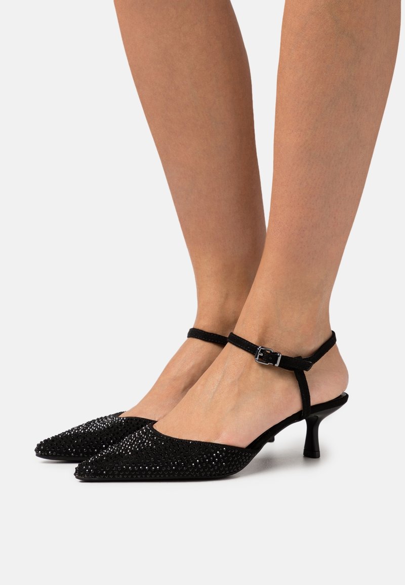Escarpins slingback noirs à bout pointu avec brides à la cheville et petits clous décoratifs noirs portés aux pieds sur un fond uni.