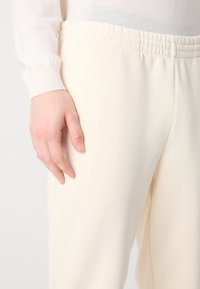 Crèmegekleurde broek met een zachte textuur en elastische tailleband. Een hand steunt op het bovenbeen, waardoor de pasvorm en de val van het kledingstuk worden benadrukt.