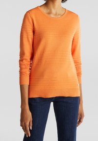Orangenes Strickpullover mit horizontalem Rippmuster, rundem Ausschnitt und langen Ärmeln, kombiniert mit dunkelblauen Jeans.