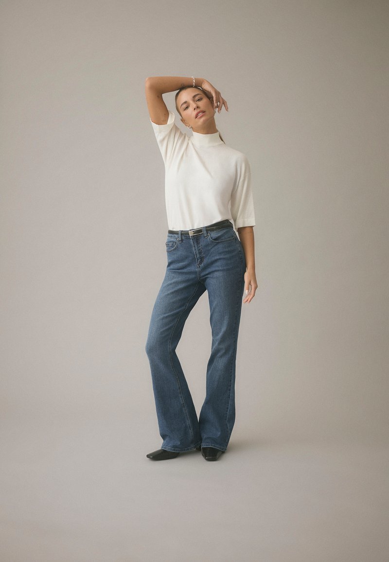 Bijela majica s kratkim rukavima i visokim ovratnikom uparena s plavim trapericama zvonolikog kroja. Kombinacija uključuje crni remen i crne cipele špic od prsta.