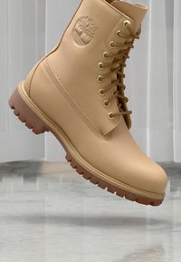 Botte en cuir cuir tan à lacets avec semelle robuste, logo d'arbre en relief sur le côté, photographiée en plein air au-dessus d'un sol poli et d'un fond à panneaux blancs.