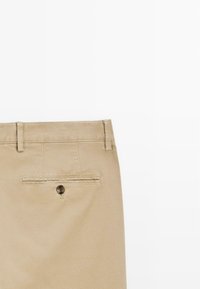 Pantalon beige vu de dos montrant des passants de ceinture et une poche passepoilée à bouton unique, sur un fond blanc uni.