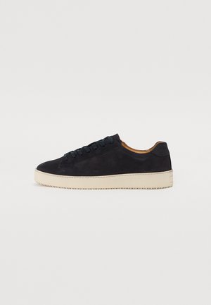Black suede sneaker with round toe, contrasting beige rubber sole, flat laces, and subtle textured details on the upper.