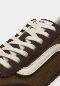 Baskets Vans marron et marron foncé avec une bande blanche et des lacets blancs, présentant un gros plan détaillé de la tige et de la languette.