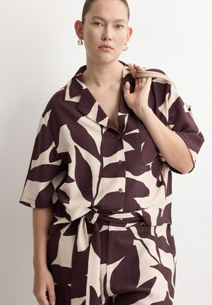 Žena odjevena u odgovarajuću široku košulju i hlače s velikim apstraktnim uzorkom smeđeg i bež lista, drži remen bež torbe preko ramena.