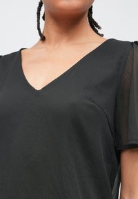 Μαύρο μπλούζα με V λαιμόκοψη, υφή στο ύφασμα, κοντά διάφανα μανίκια και άνετη εφαρμογή. Ξεχωρίζει για τη λεία ροή και τον μινιμαλιστικό σχεδιασμό της.