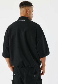 Camicia nera oversize realizzata in tessuto testurizzato, con maniche corte, orlo arrotondato e un piccolo logo sul retro.