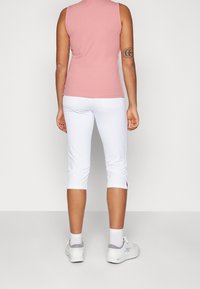 Persona de pie de espaldas, vestida con una camiseta sin mangas rosa, pantalones cortos blancos, calcetines blancos y zapatillas blancas con ribete gris.