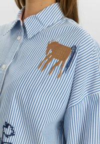 Gestreifte Bluse in Hellblau und Weiß, mit brauner Pferdestickerei und dunkelblauen Akzenten. Hergestellt aus Baumwolle mit klassischem Kragen.