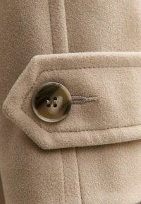 Beżowa tkanina z wełny na płaszcz, z dużym, okrągłym, marmurkowym guzikiem z dwoma otworami, przymocowanym dopasowaną nicią i widocznym akcentem szwu.