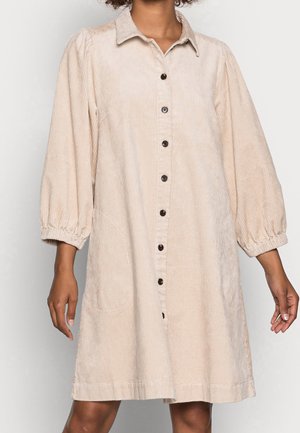 Femme portant une robe-chemise en velours côtelé beige avec des boutons noirs et des manches bouffantes trois-quarts.