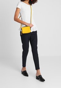 Top blanco de manga corta, pantalones ajustados slim-fit a rayas negras, mules negras y un pequeño bolso cruzado amarillo con cierre de cremallera y correa ajustable.