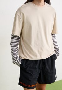 Person med en beige kortärmad skjorta över en grå och vit randig långärmad tröja, iförd svarta Nike Athletics shorts, med händerna i fickorna.