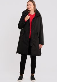 Cappotto impermeabile nero con colletto alto, chiusura a bottoni e tasche laterali, indossato sopra un maglione rosso e pantaloni neri.