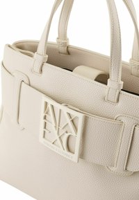 Borsa a mano in pelle crema con superficie texturizzata, doppi manici e una chiusura con logo in evidenza. Forma rettangolare con un design strutturato.