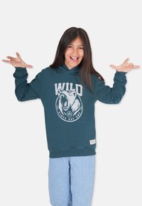 Teal hoodie med en björngrafik, texten "WILD" och "SINCE DAY ONE" nedanför. Har en känguruficka och ribbade muddar. Matchas med ljusa jeansshorts.