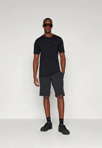 Μαύρη κοντομάνικη μπλούζα σε συνδυασμό με σκούρες γκρι σορτς. Το μοντέλο φοράει σκούρα γυαλιά ηλίου και μαύρα αθλητικά παπούτσια, αναδεικνύοντας μια ανάλαφρη αθλητική εμφάνιση.