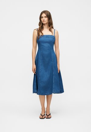 Jonge vrouw met lang bruin haar, gekleed in een mouwloze blauwe denim midi-jurk en zwarte open teen sandalen, staand tegen een effen achtergrond.
